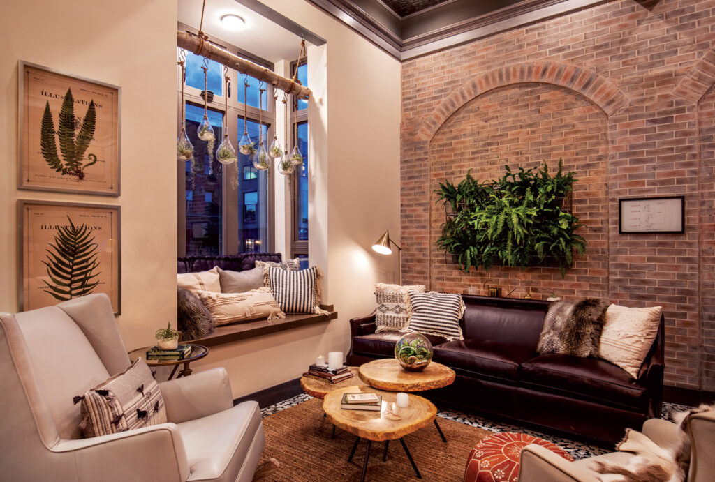 Cozy living room with a brown leather sofa, white armchair, circular wood tables, brick accent wall, indoor plants, and large window with hanging glass bulbs.