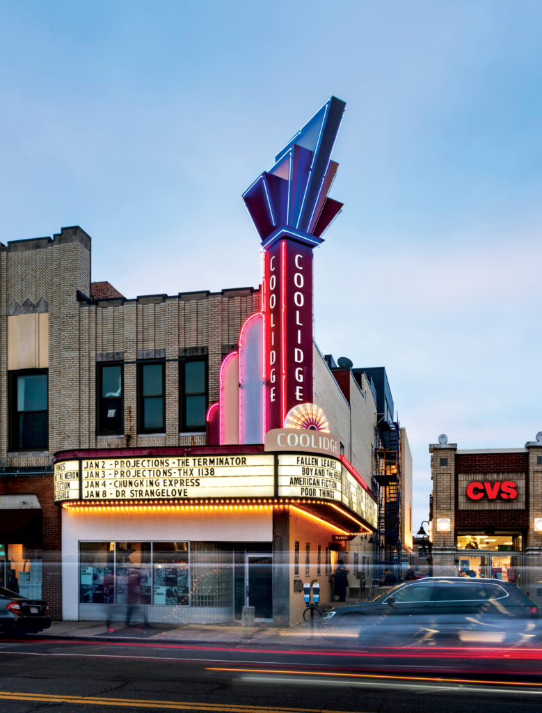 The Coolidge Corner Theatre with neon lights and marquee displaying movie titles, located next to a CVS Pharmacy on a city street at dusk.