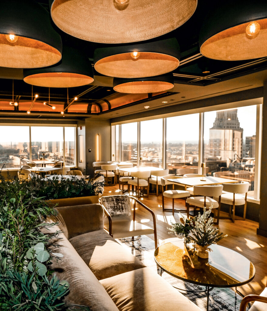 Modern lounge with large windows, city views, beige seating, round tables, potted plants, and ceiling lights in daylight.