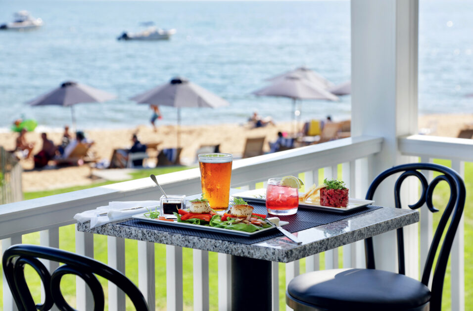 A table with two cocktails and plates of food overlooks a beach with people, umbrellas, and boats on the water in the background.