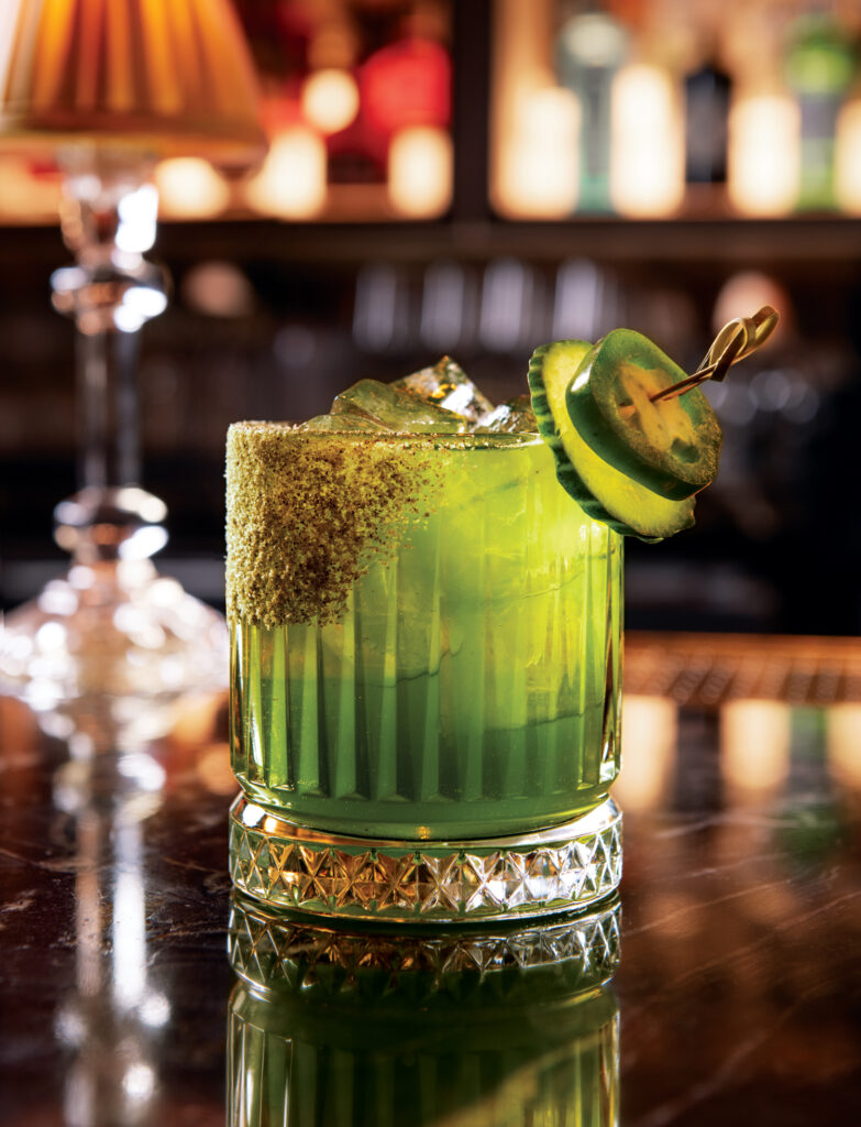 A green cocktail garnished with a jalapeño slice and a salted rim sits on a polished bar counter, with a lamp and blurred bottles in the background.