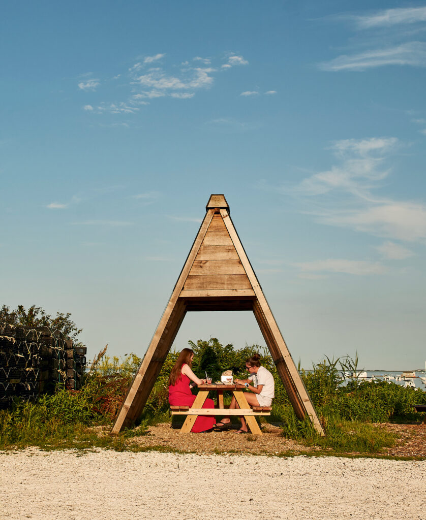Best Things to Do in Massachusetts This Summer. Three people sit at a picnic table under a wooden triangular shelter outdoors on a sunny day, with greenery and sky in the background.