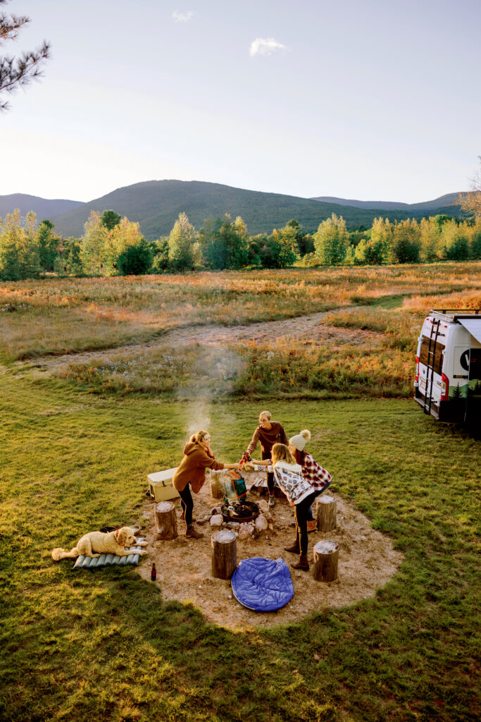 Best Things to Do in Massachusetts This Summer. Two people cook over a campfire surrounded by tree stumps, with a dog lying nearby and a van parked in a grassy field with mountains and trees in the background.