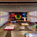 Large gallery space with colorful textile art installations, patterned illuminated panels on the floor, and a vibrant abstract mural covering the back wall.