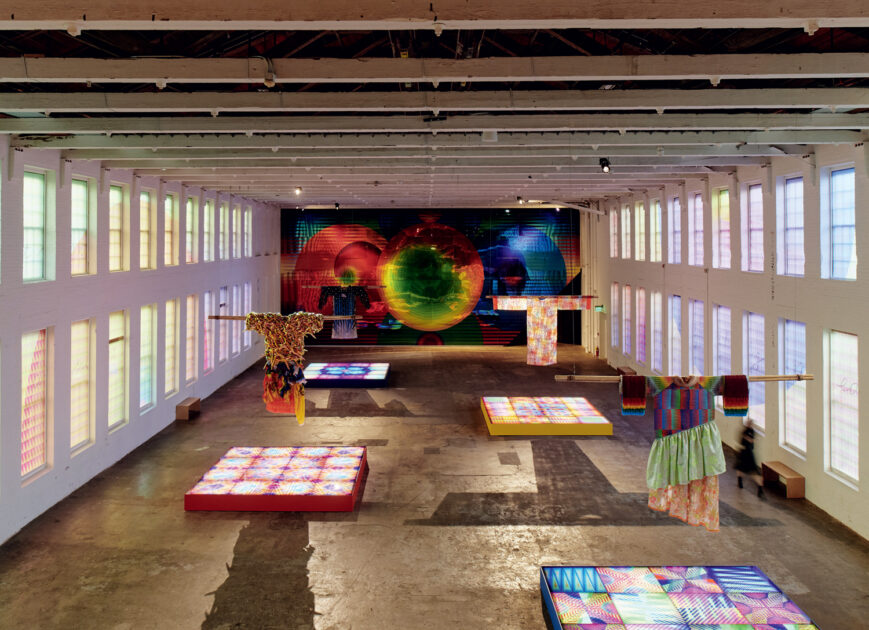 Large gallery space with colorful textile art installations, patterned illuminated panels on the floor, and a vibrant abstract mural covering the back wall.