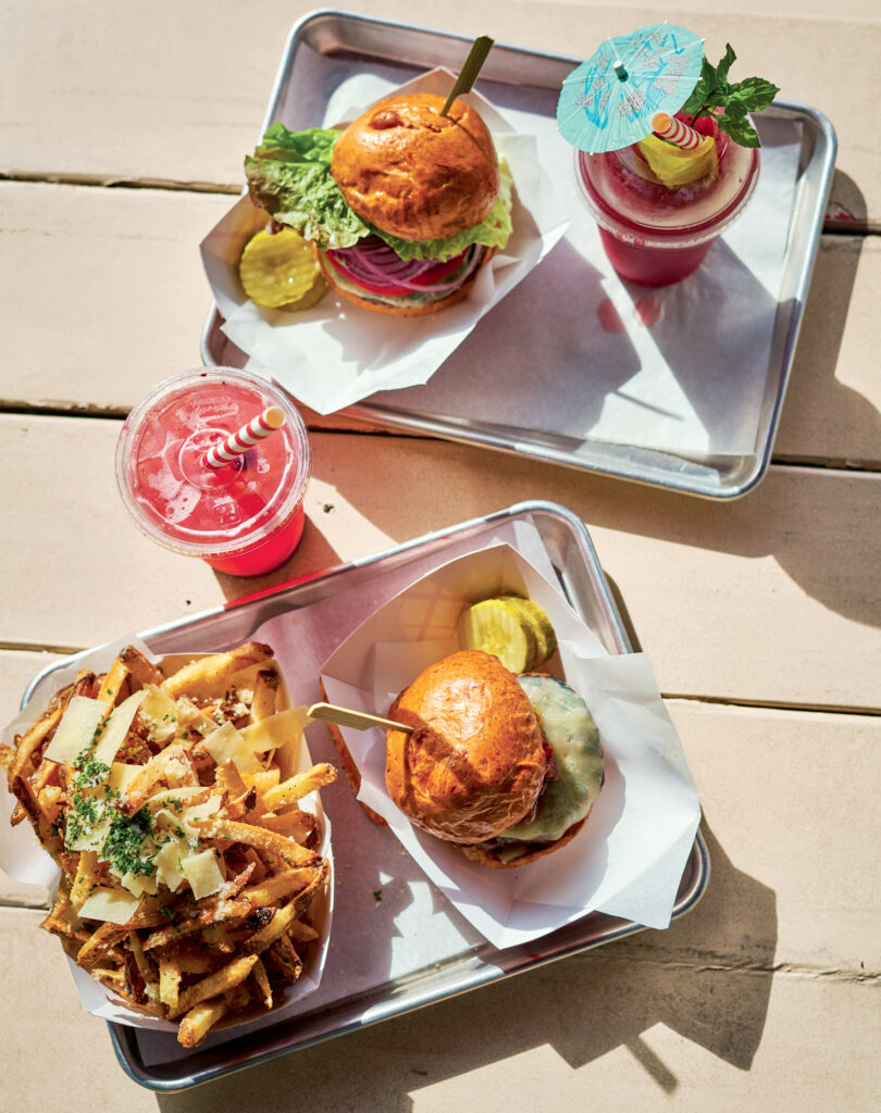 Two trays with cheeseburgers, pickles, fries topped with cheese, and two pink drinks garnished with umbrellas and citrus slices on a light wooden table.
