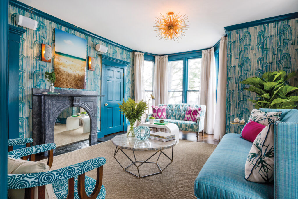 Vibrant living room with patterned blue wallpaper, teal accents, modern furniture, a marble fireplace, geometric coffee table, and natural light from large windows.