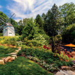 A scenic garden landscape with a windmill on the left, stone steps, lush greenery, and people gathered near a table with an orange umbrella on the right.