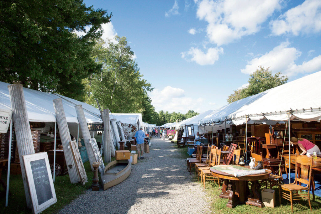 Outdoor flea market scene with white tents on both sides of a gravel path, displaying various furniture and antique items under a blue sky with scattered clouds.