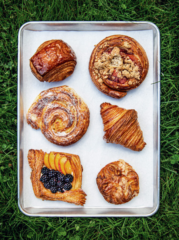 10 Best Things to Do in Maine This Summer. A metal tray with six assorted pastries, including a fruit danish, croissant, cinnamon roll, and other baked goods, sits on parchment paper on green grass.