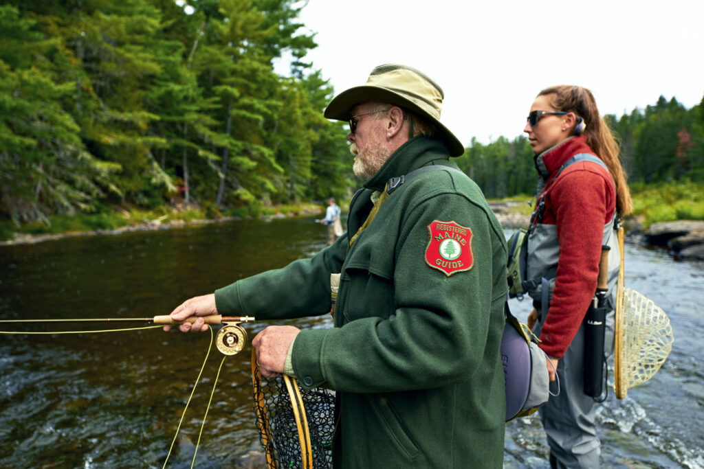 An older man in a ranger uniform and a young woman, both fly fishing in a forested river, stand near the water's edge with fishing gear.
