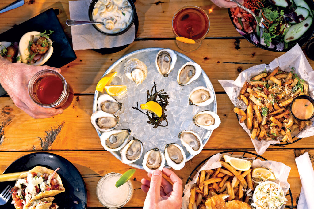 A table with plates of oysters on ice, fries, tacos, salads, and cocktails, with hands reaching for food on a wooden surface.