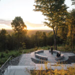 A circular stone seating area overlooks a forested landscape at sunset, with two people sitting on the benches surrounded by tall trees. 2025 Maine Travel Guide | Hotels, Dining & Attractions