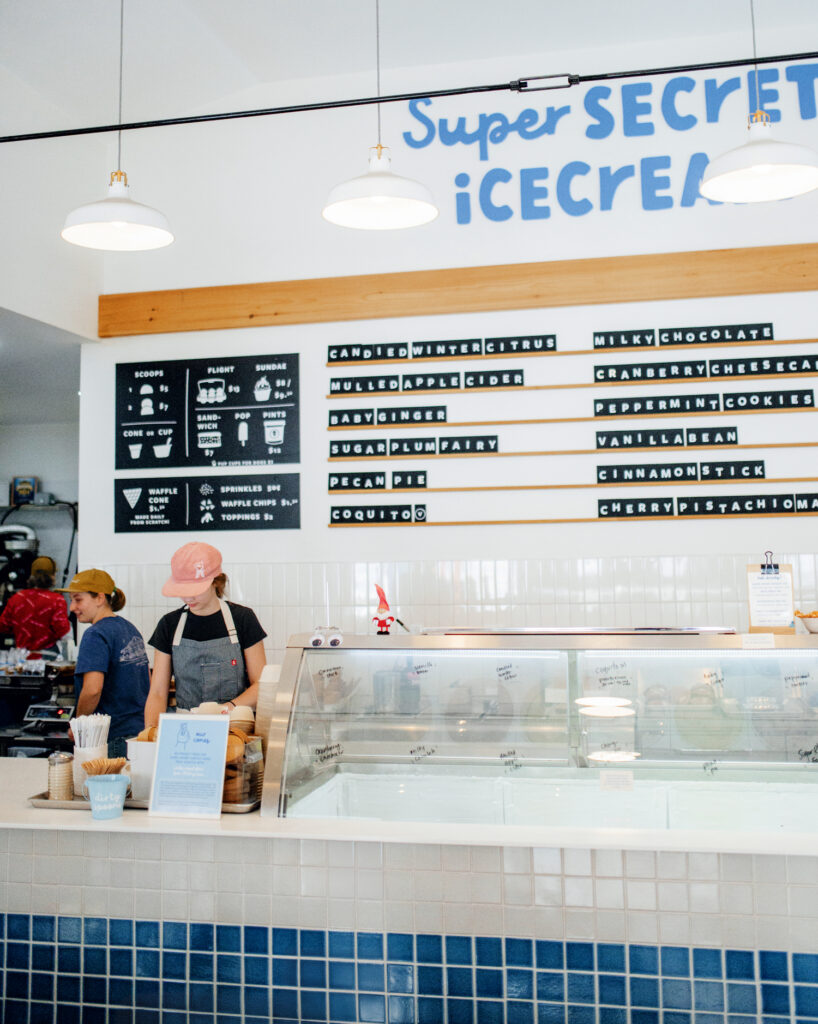 10 Best Things to Do in New Hampshire This Summer 2025. Two employees work behind the counter of an ice cream shop with a menu board listing ice cream flavors and a sign that reads "Super Secret Ice Cream" on the wall.