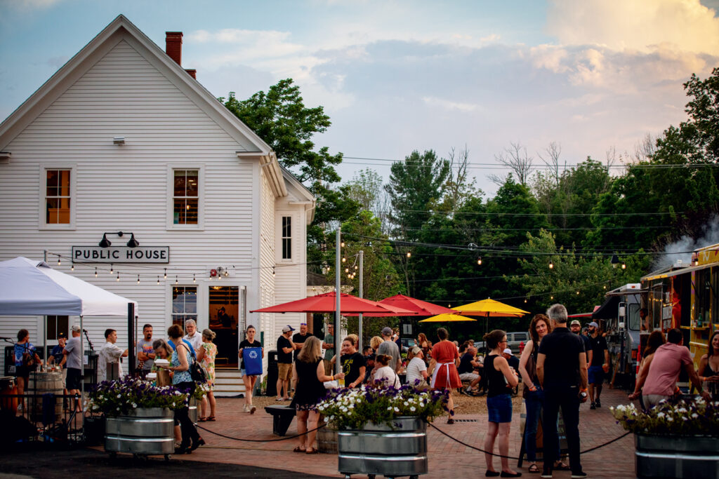 10 Best Things to Do in New Hampshire This Summer 2025. People gather outdoors near a white building labeled "Public House," with food trucks, tables, string lights, and trees in the background.