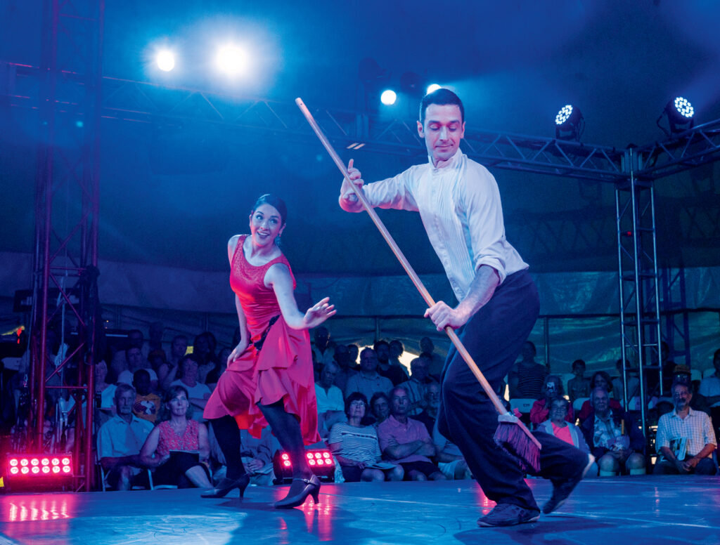 10 Best Things to Do in New Hampshire This Summer 2025. A man and woman perform a dance routine on stage in front of an audience; the man holds a broom while both wear formal attire under stage lighting.