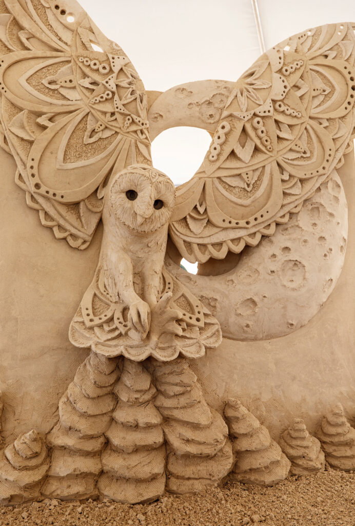 Best Things to Do in Rhode Island This Summer. Intricate sand sculpture of an owl with ornate wings, perched in front of a large moon and surrounded by cone-shaped sand formations.