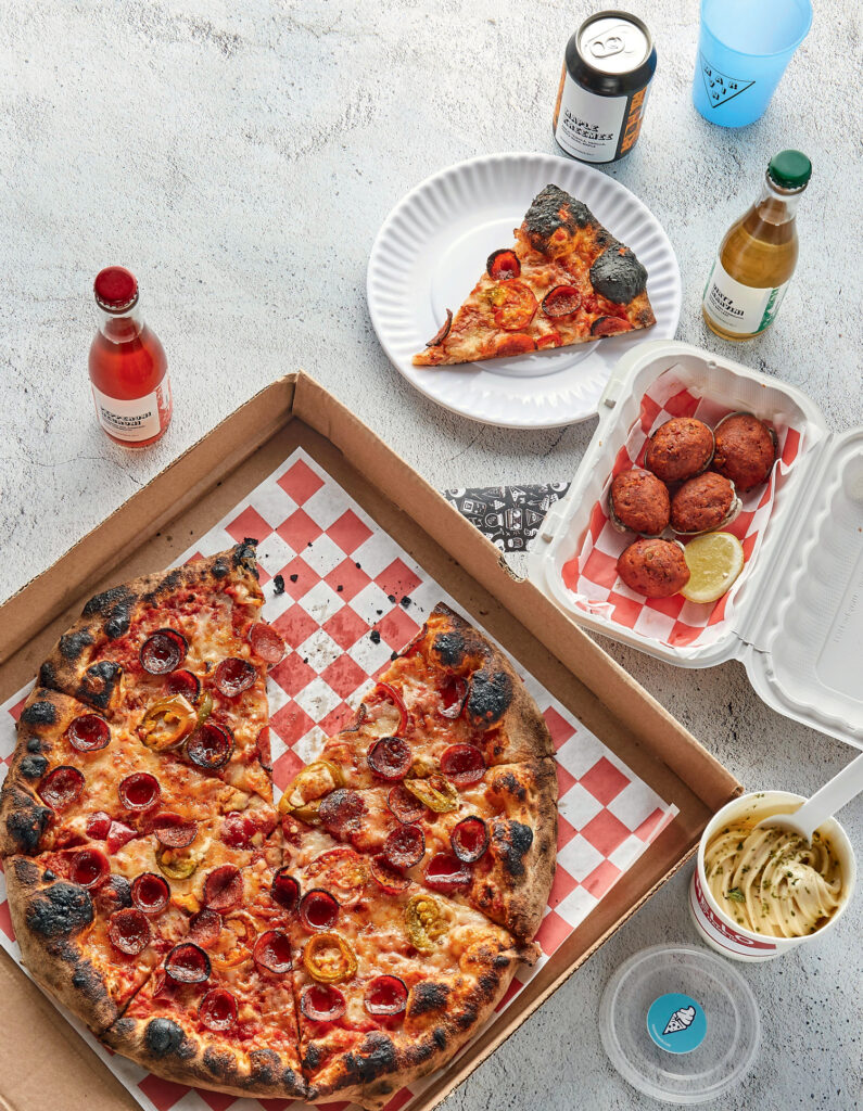 Best Things to Do in Rhode Island This Summer. A pepperoni pizza in an open box, a slice on a plate, arancini balls in a takeout container, drinks, and a cup of gelato on a light textured surface.