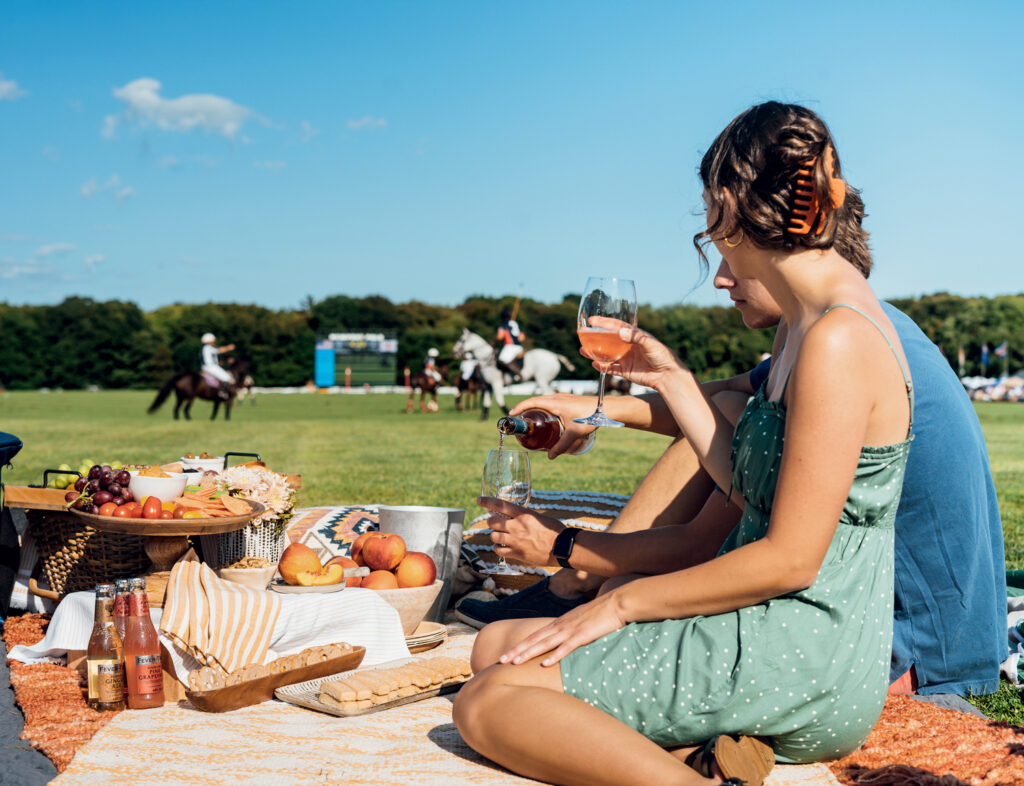 Two people sit on a picnic blanket with food and drinks, watching a polo match on a grassy field under a clear blue sky.