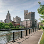A wooden boardwalk runs beside a river, with modern and historic buildings in the background under a partly cloudy sky.