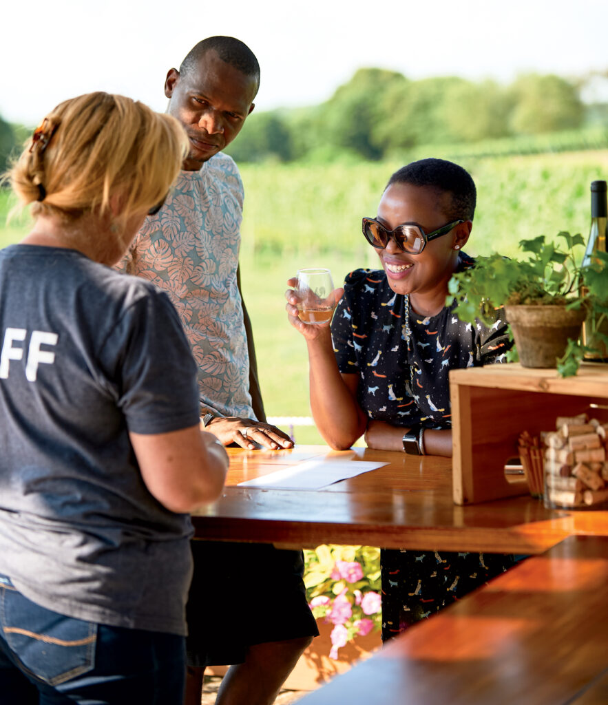 Two people stand at an outdoor counter, interacting with a staff member. One person holds a glass and smiles. There are wine bottles, corks, and plants on the counter.