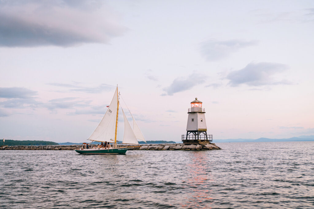 Best Things to Do in Vermont This Summer. A sailboat passes by a small lighthouse on a rocky breakwater at sunset, with calm water and a partly cloudy sky in the background.