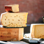 Best Things to Do in Vermont This Summer. Large blocks of cheese, one with holes, are stacked on a wooden box and table, with small cubes of cheese in front, displayed for sampling.