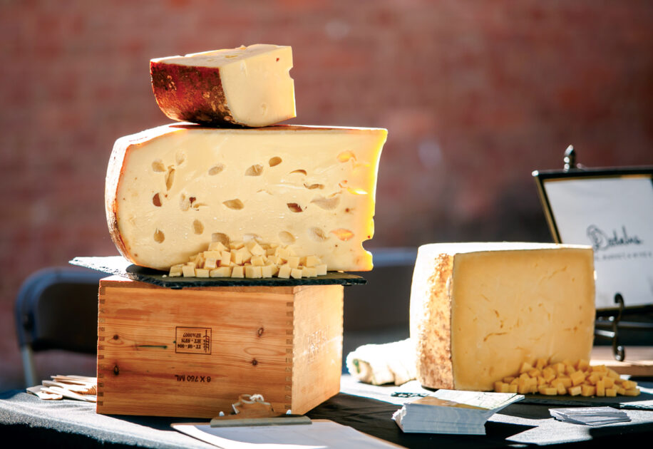 Best Things to Do in Vermont This Summer. Large blocks of cheese, one with holes, are stacked on a wooden box and table, with small cubes of cheese in front, displayed for sampling.