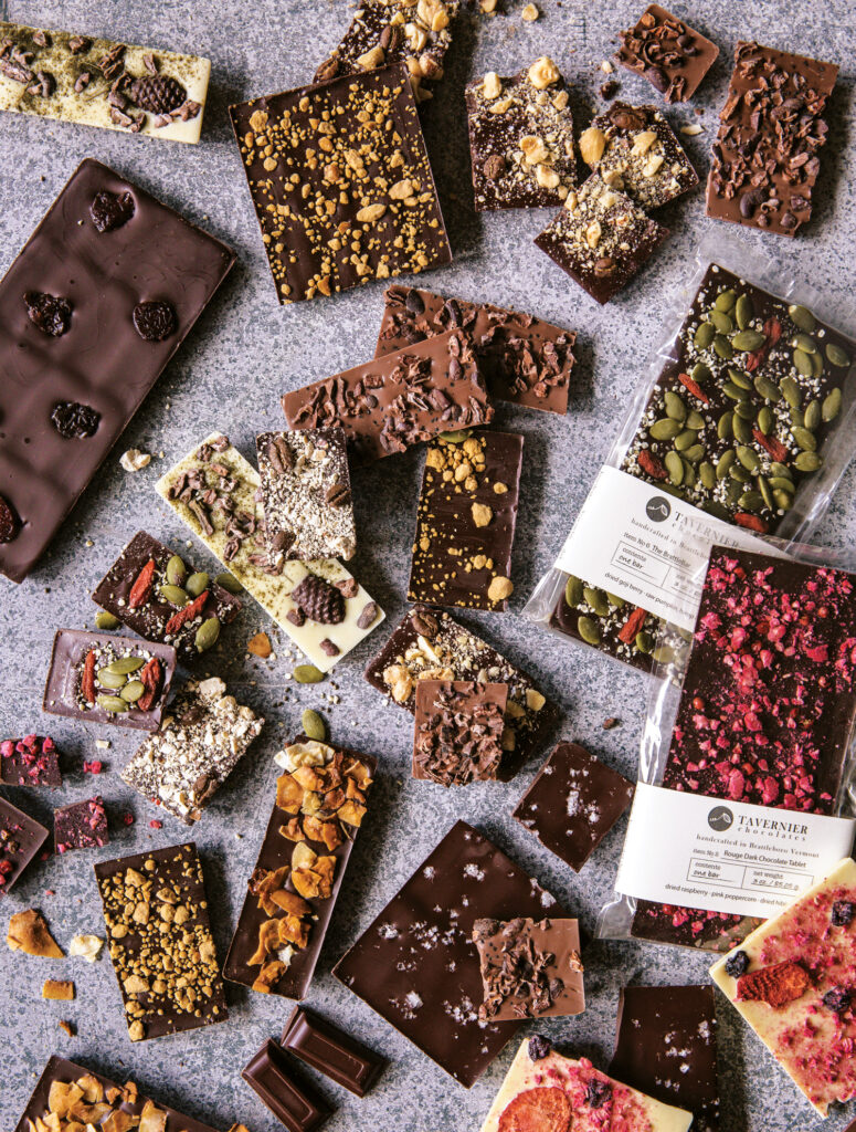 An assortment of chocolate bars and pieces with various toppings, including nuts, seeds, berries, and dried fruit, arranged on a gray surface.