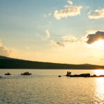People paddleboarding, kayaking, and standing on rocks by the lake during sunset, with hills in the background and scattered clouds in the sky.