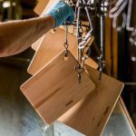 A hand in a blue glove holds a wooden board with hooks in a drying line with several similar boards hanging. Water droplets are visible on the surface.