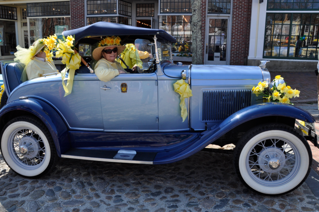Blue vintage car decorated with yellow flowers and ribbons, carrying passengers wearing yellow hats and driving on a cobblestone street.