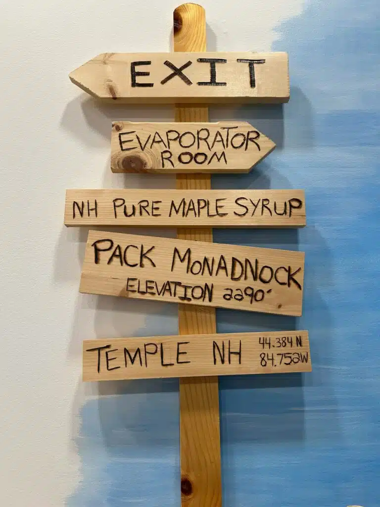 Wooden signs on a post indicating directions: Exit, Evaporator Room, NH Pure Maple Syrup, Pack Monadnock at elevation 2290 feet, and Temple NH with coordinates. Background is blue and white.