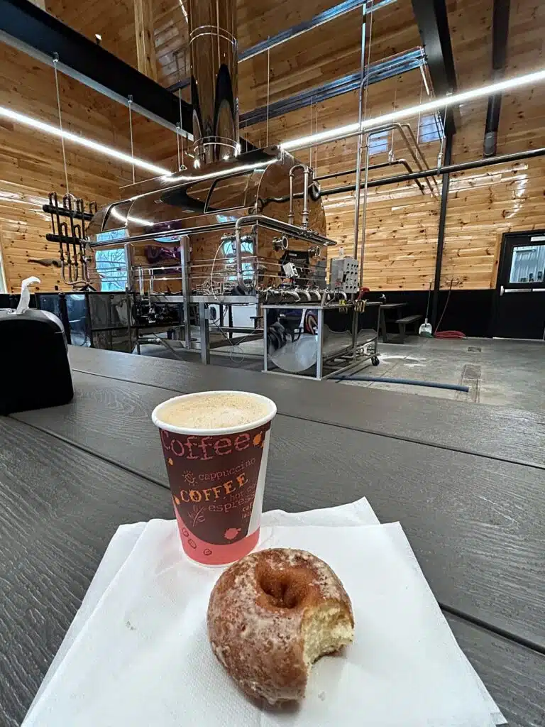 Cup of coffee and half-eaten donut on a napkin on a wooden table inside a modern building with a large espresso machine in the background.