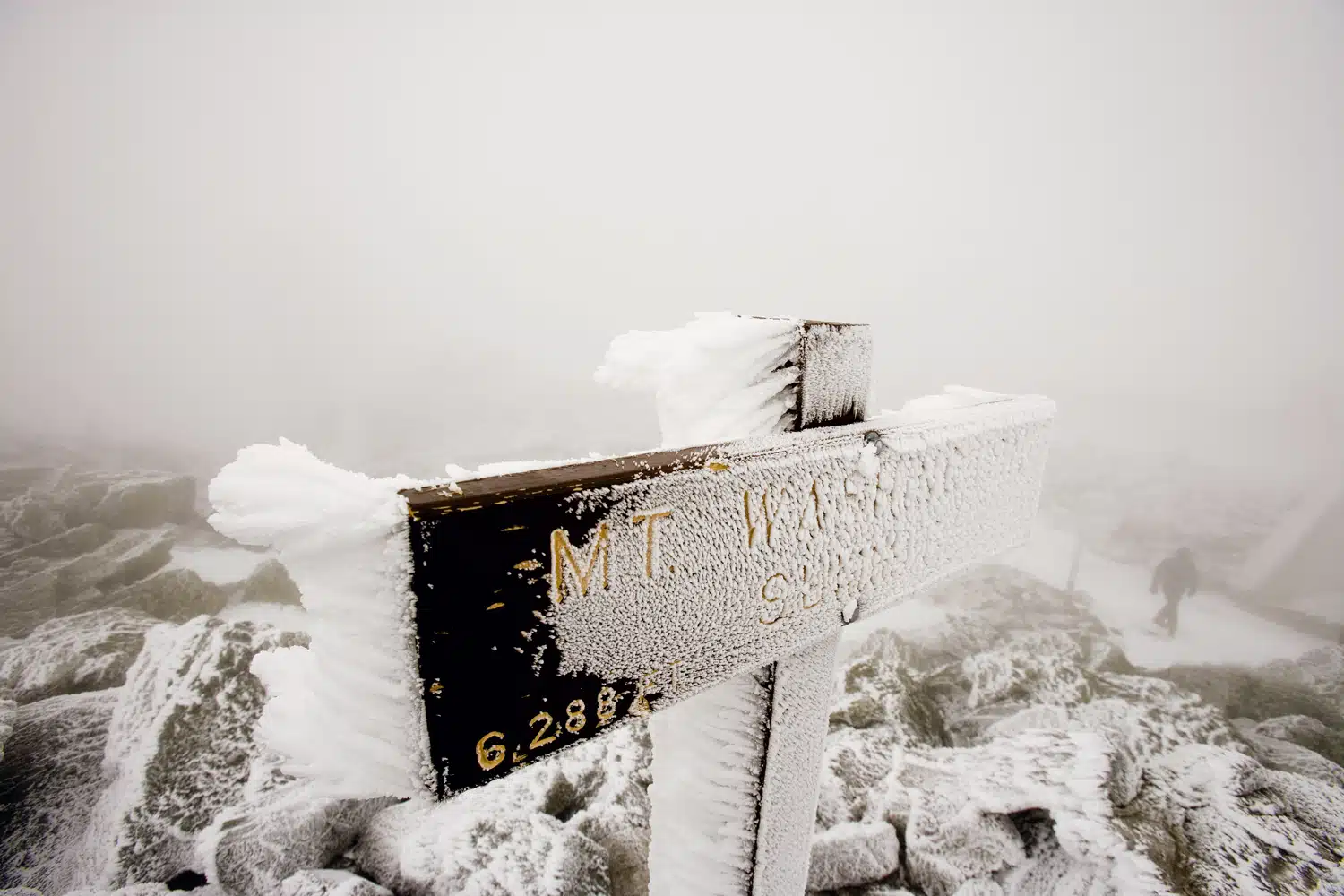 Snow-covered wooden sign at Mt. Washington summit, partially obscured by fog, with rocks and a person in the background.
