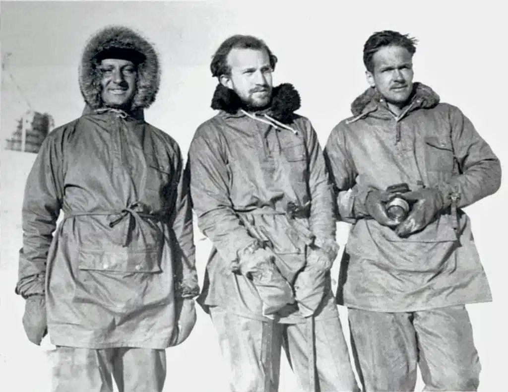 Three explorers in fur-lined coats stand together in snowy conditions, wearing gloves and warm clothing, with one holding a camera.