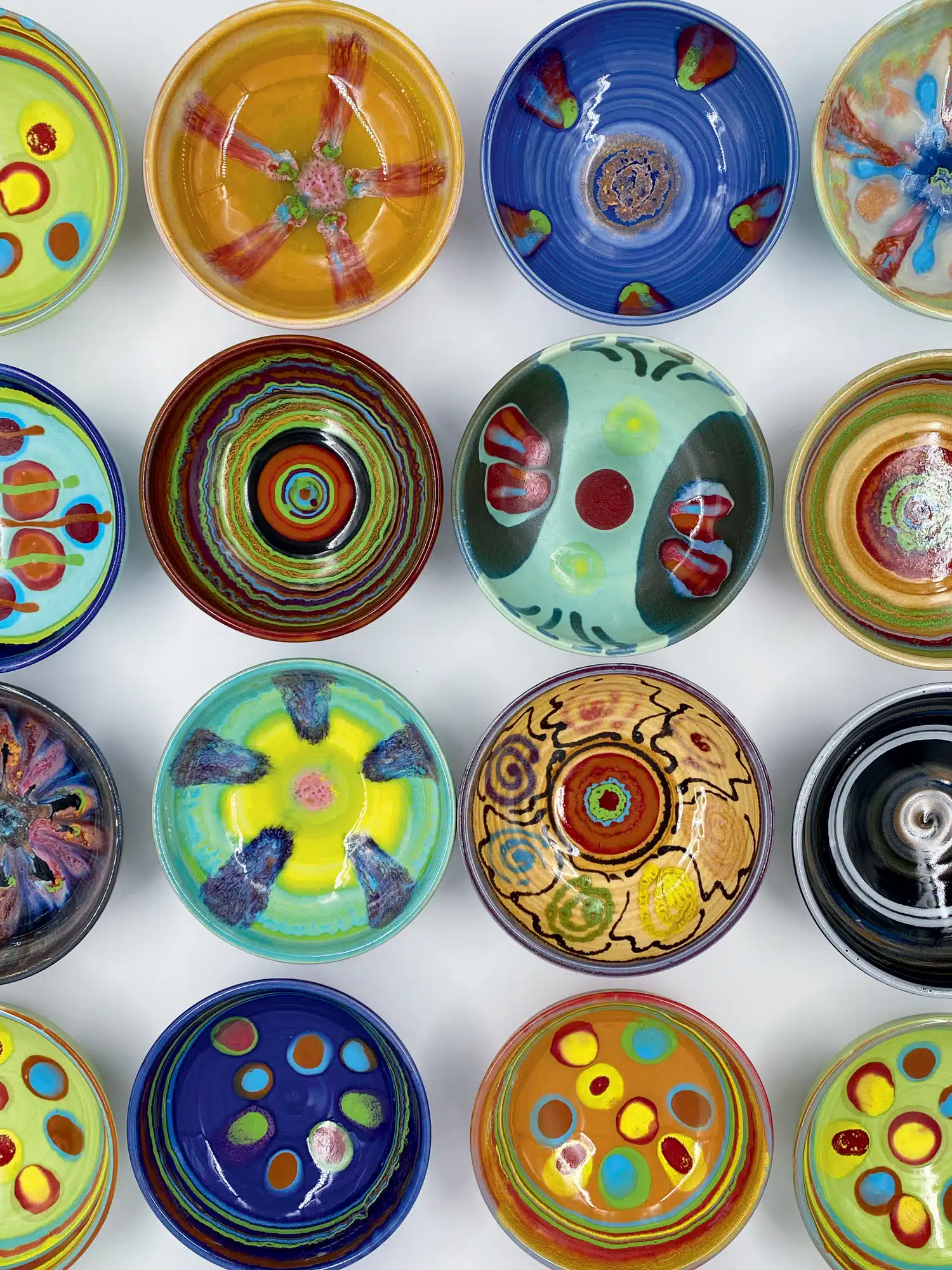 A collection of sixteen colorful, hand-painted ceramic bowls with various abstract patterns, arranged in a grid on a white background.