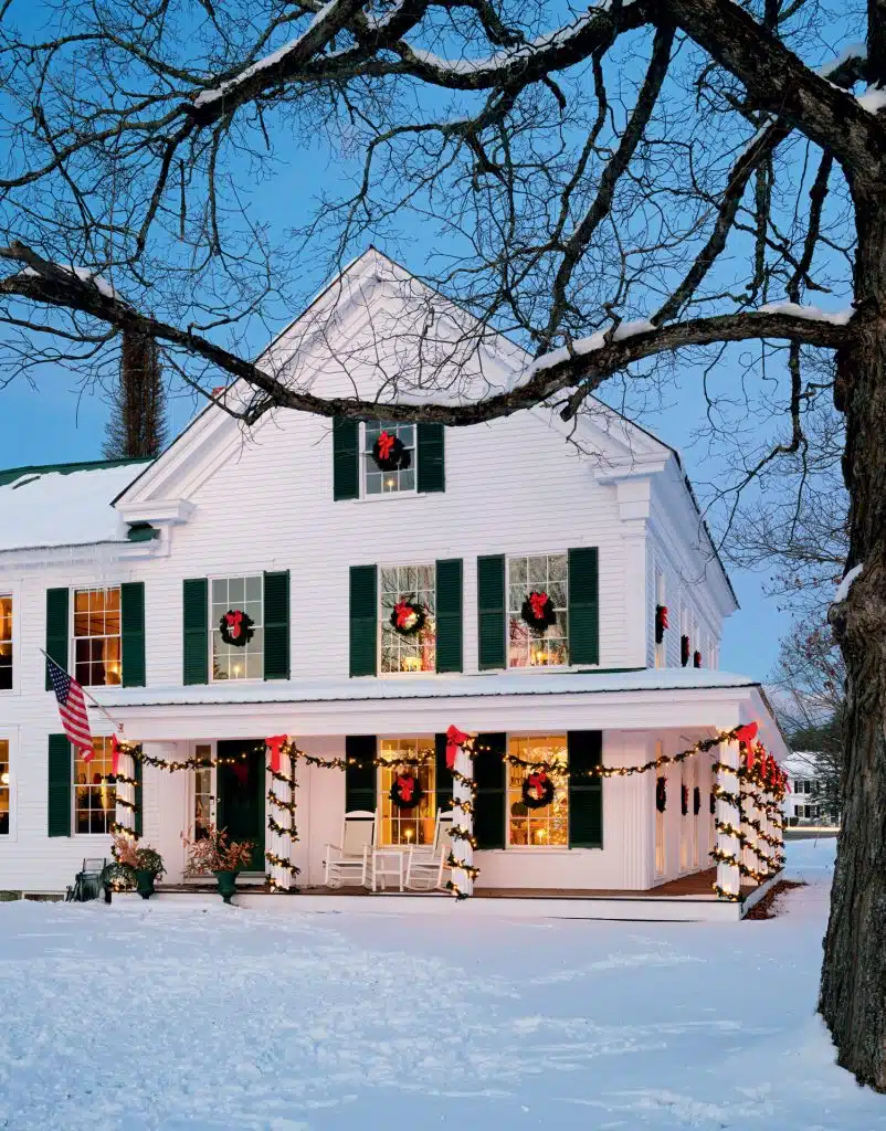 A white house with green shutters is decorated with wreaths and lights. Snow covers the ground, and a bare tree branches in the foreground.