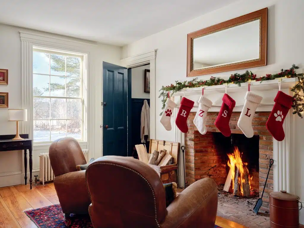 A cozy living room with a lit fireplace, leather armchairs, and stockings hanging on the mantel. A window shows a snowy landscape outside.