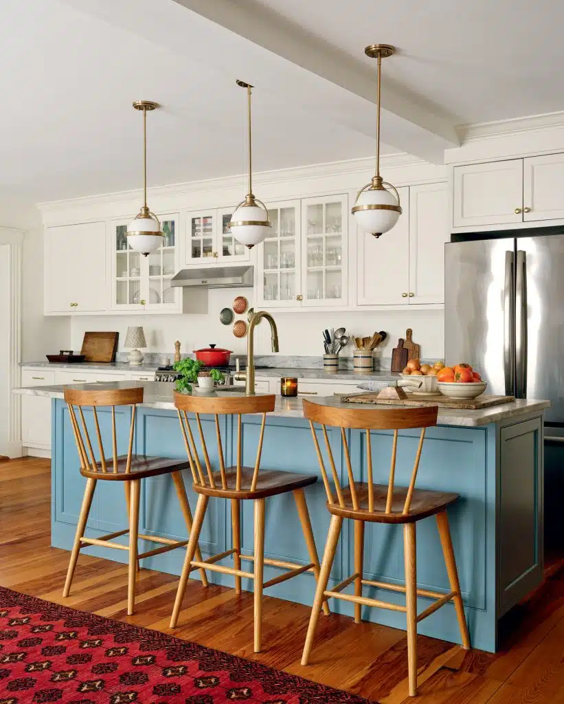 A kitchen with a blue island, three wooden stools, pendant lights, white cabinets, and stainless steel appliances. A red rug decorates the wooden floor.