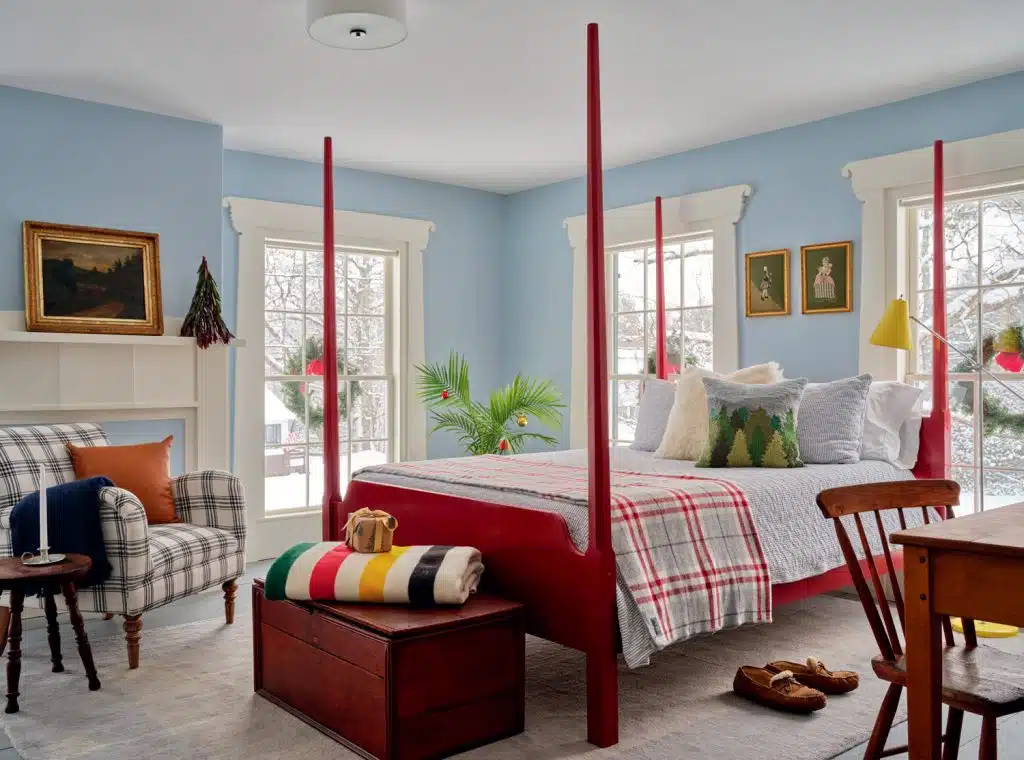 Cozy bedroom with a four-poster red bed, plaid bedding, a checked armchair, and a wooden chest. Blue walls feature artwork; large windows bring in natural light.