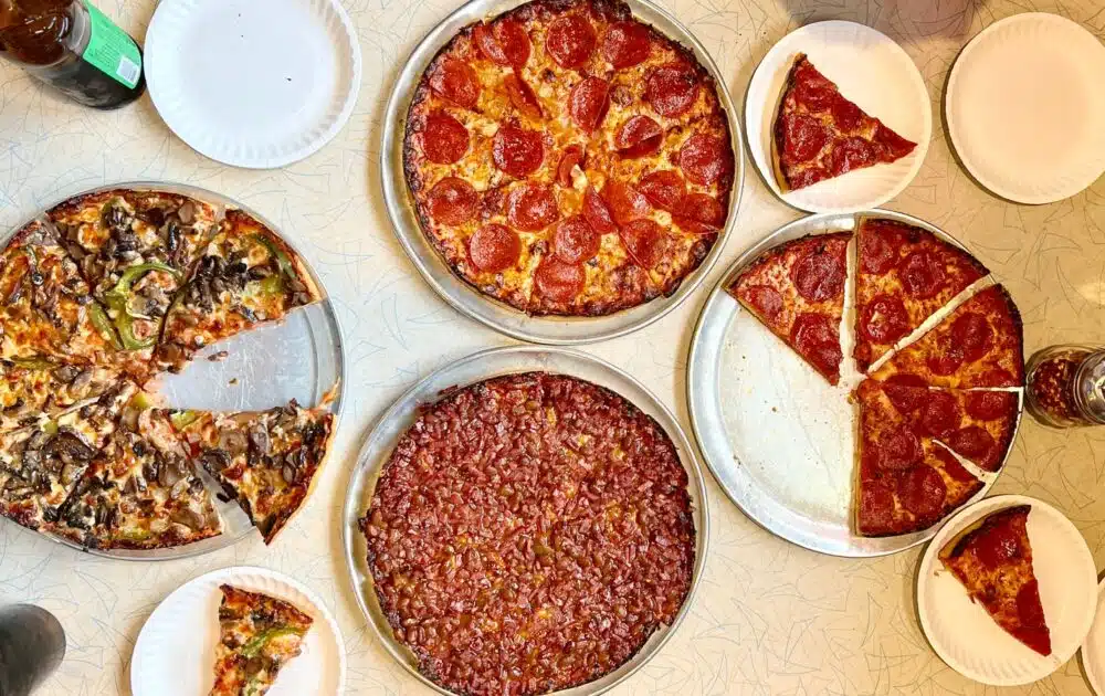 Five pizzas on trays with various toppings, including pepperoni and vegetables, are placed on a table with paper plates and drinks.