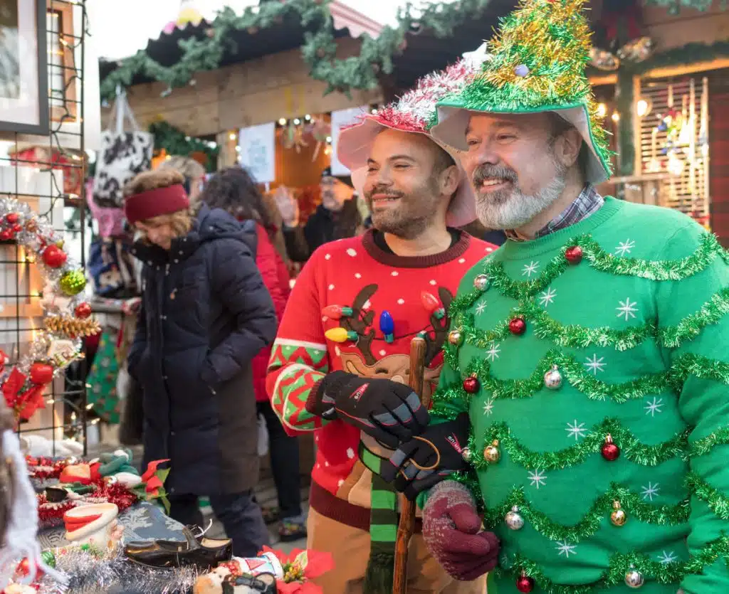 Two people in festive attire and decorated hats stand in front of a holiday market stall with wreaths and ornaments.
