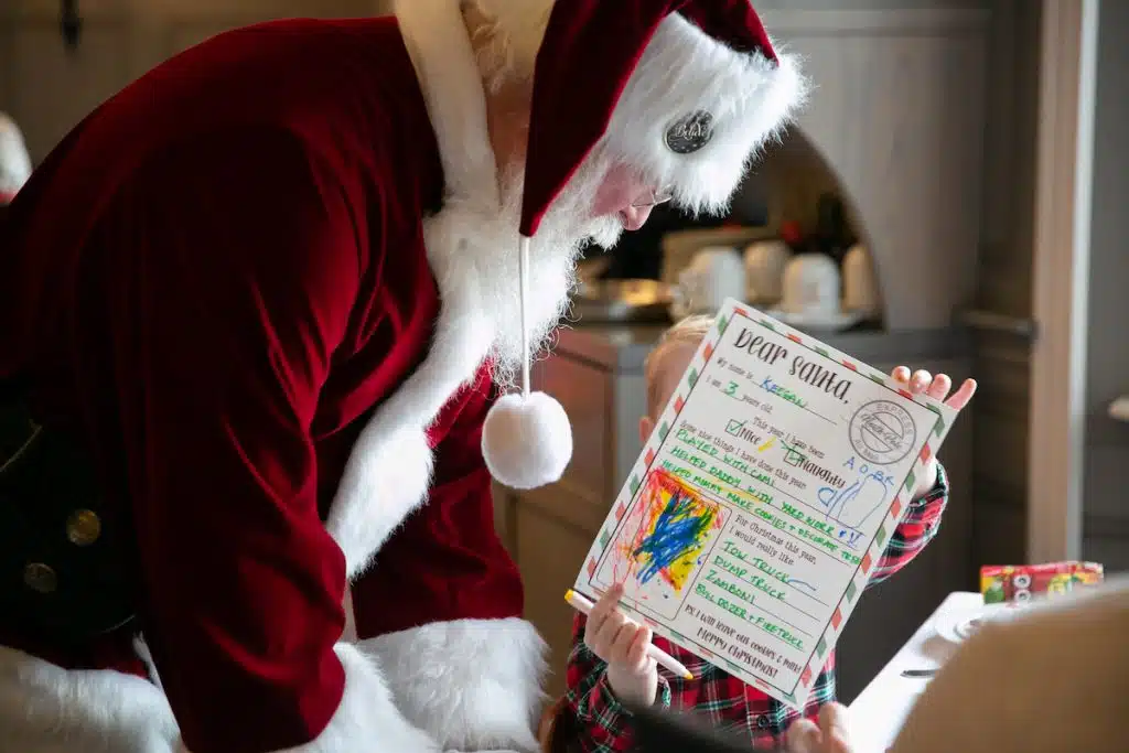 A person dressed as Santa Claus looks at a child holding a colorful letter addressed to Santa.