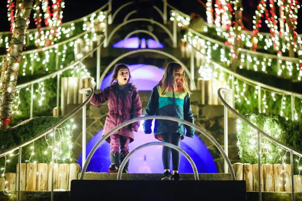 Two children walk down illuminated steps surrounded by festive lights at night.