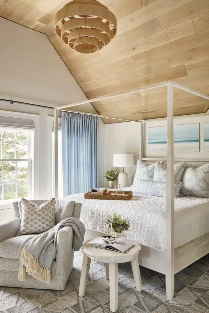 Cozy bedroom with a four-poster bed, light wood ceiling, and a window with blue curtains. A plush armchair and side table with books and flowers complete the decor.