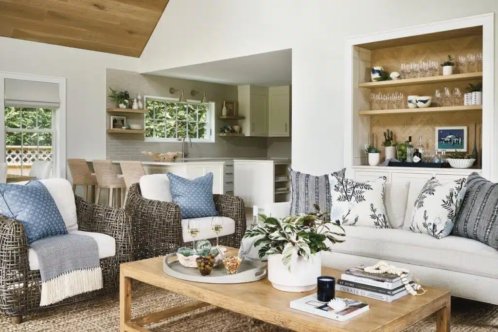 Nantucket Looms - A modern living room with a sofa, wicker chairs, a wooden coffee table, and decorative plants. The open kitchen and dining area feature a breakfast bar with stools and a built-in shelving unit.