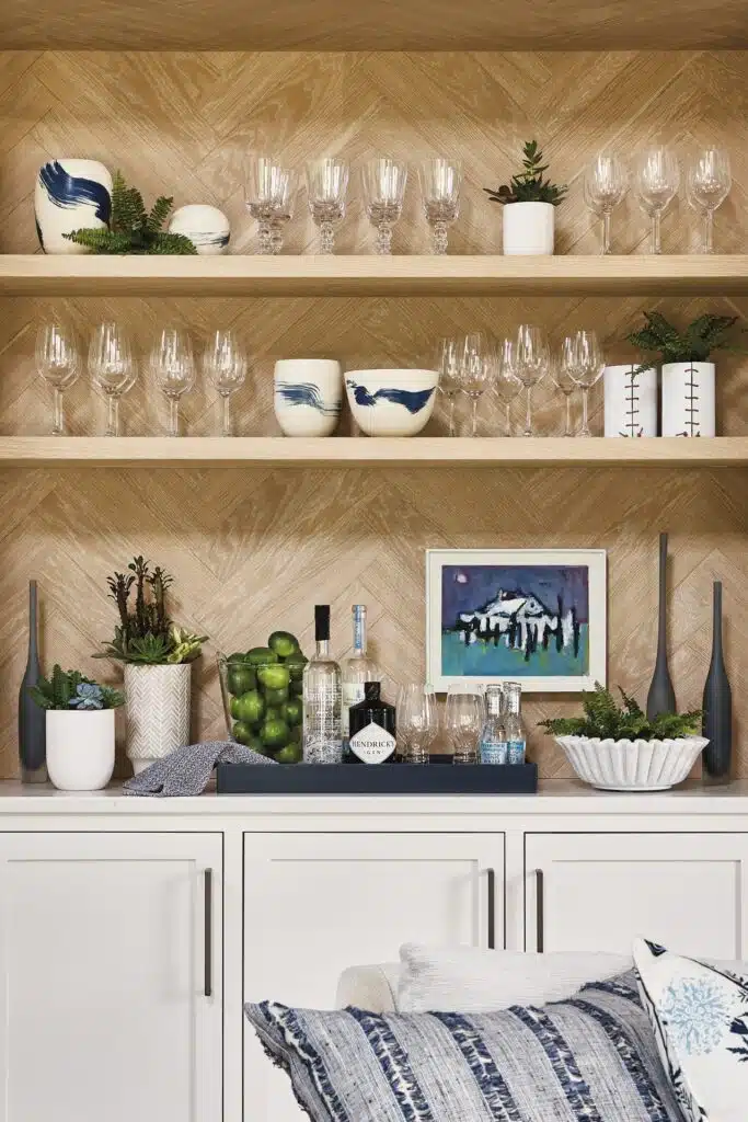 Nantucket Looms - Shelving unit with wine glasses, decorative vases, and plants. Below, a counter displays bottles, limes, and a framed picture. White cabinetry beneath. Cushions in the foreground.
