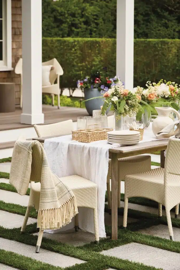 Outdoor dining table with woven chairs, a white tablecloth, and a centerpiece of flowers and greenery. Plates and a woven basket are on the table, set in a garden with a hedge background.