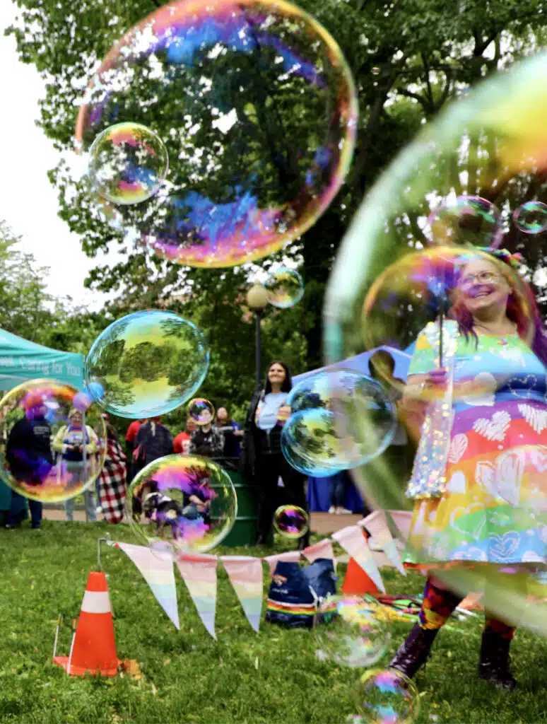 A person in colorful clothing stands on grass, surrounded by large soap bubbles, with people and trees in the background at an outdoor event.